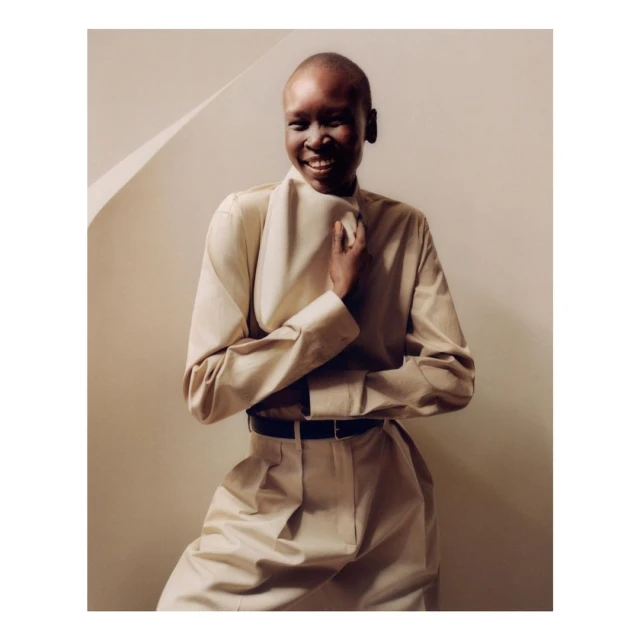 A smiling, bald, Black woman in a beige collared shirt and matching pants poses indoors against a beige wall and geometric shadow.