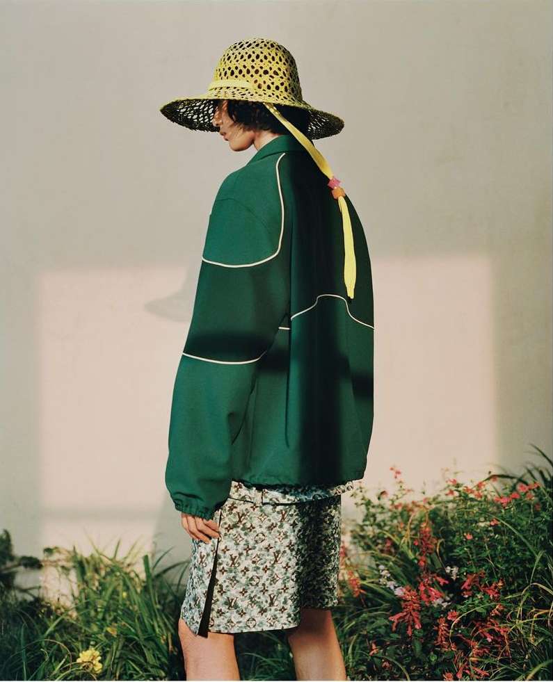 Fashion shot of a person in a green jacket, floral skirt, and straw hat posing in a studio with green plants and a minimalist backdrop.