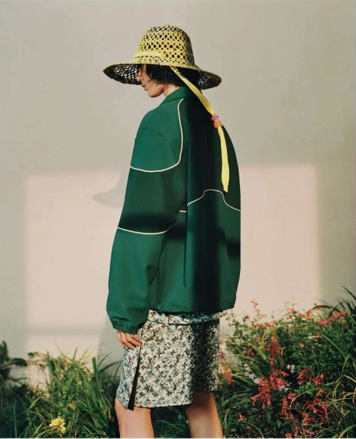Fashion shot of a person in a green jacket, floral skirt, and straw hat posing in a studio with green plants and a minimalist backdrop.