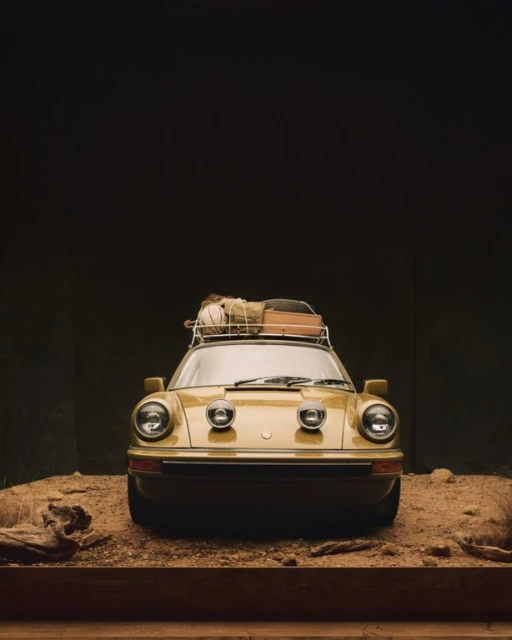 Vintage yellow Porsche 911 with luggage on the roof rack, shot in a studio against a dark background with sandy ground.