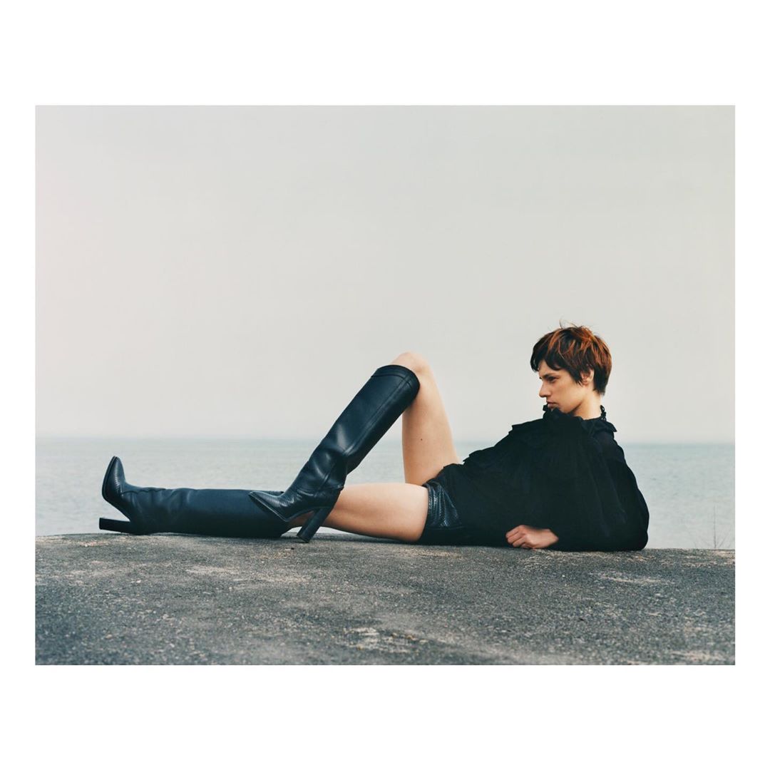 Model in black top and leather boots reclining on a concrete ledge by the water, photographed by Andrew Jacobs.