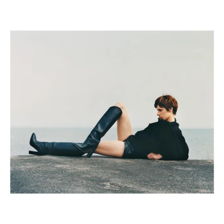 Model in black top and leather boots reclining on a concrete ledge by the water, photographed by Andrew Jacobs.