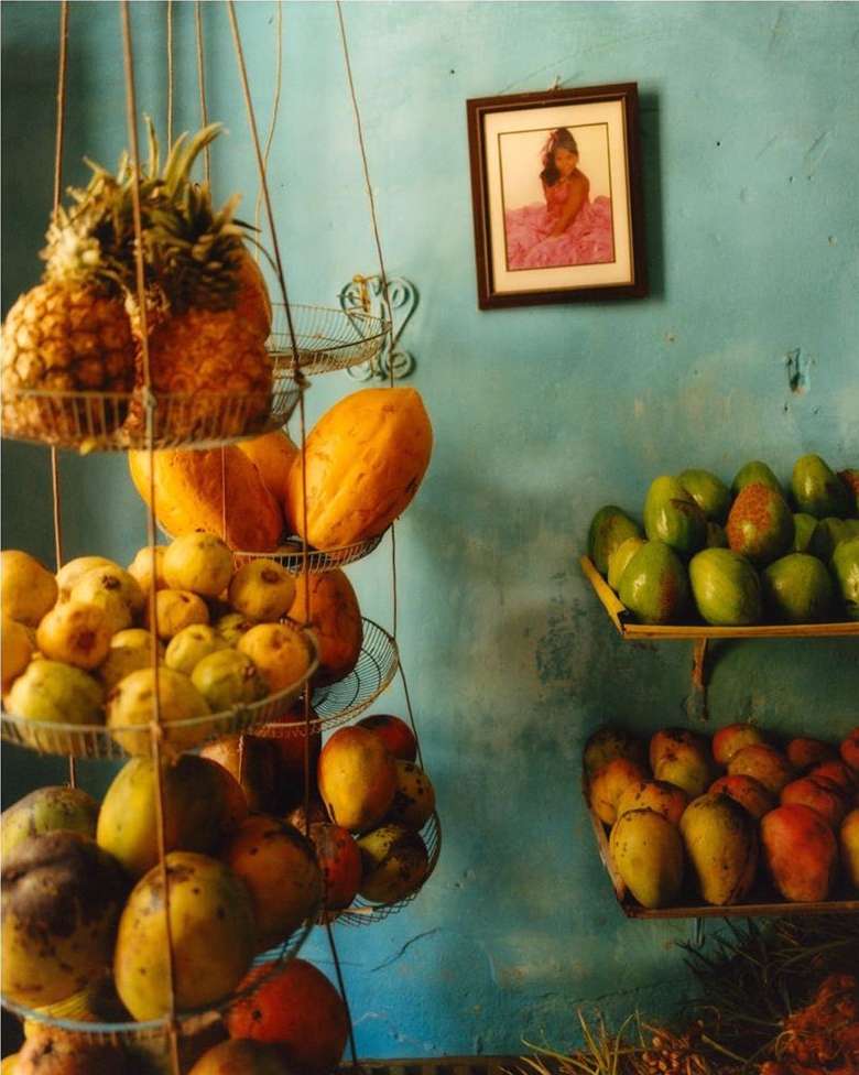 Still life of fruit for sale displayed on metal tiers, set against a blue wall in an indoor setting, enhanced with a framed image.