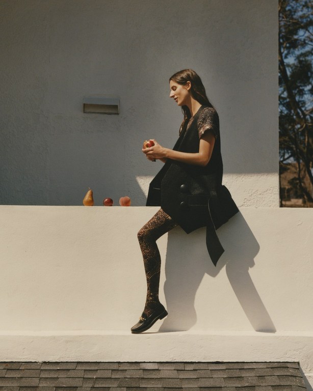 A woman in a black blazer and patterned tights sits outside on a ledge holding a red apple with fruit in the background.