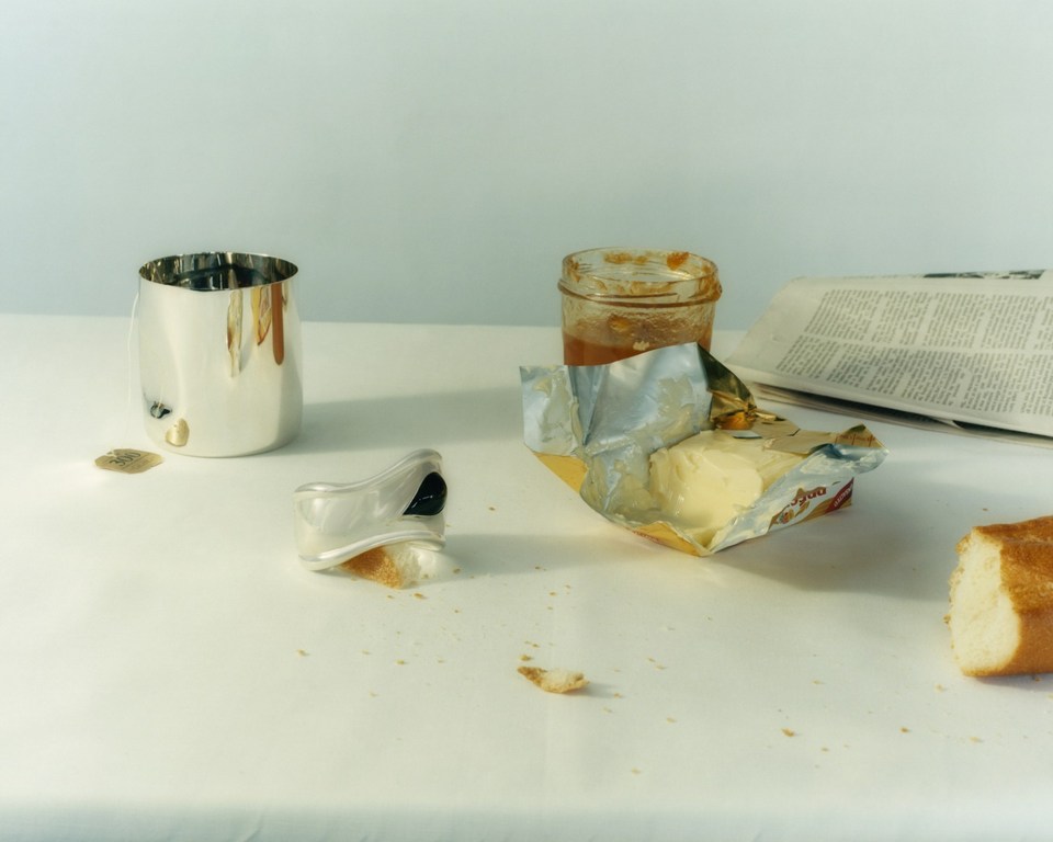 Still life photo of breakfast food including butter, baguette, and jam alongside a silver cup and newspaper, arranged on a white surface.