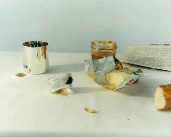 Still life photo of breakfast food including butter, baguette, and jam alongside a silver cup and newspaper, arranged on a white surface.