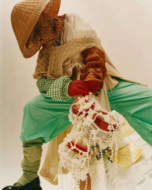 Model wearing layers of textured clothing, beaded jewelry, and conical hat in a fashion editorial shot against a white background.