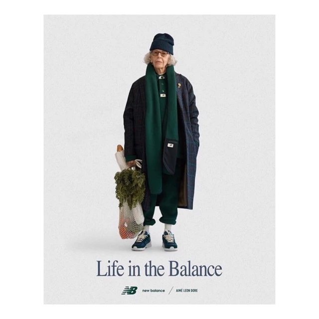 Elderly woman posing in a coat, scarf, beanie, and athletic shoes, holding a grocery bag, in a studio shot for a New Balance ad.