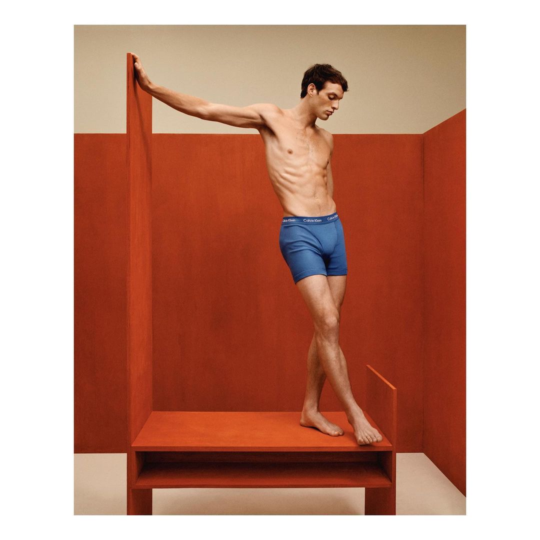 An adult male model poses indoors wearing blue underwear against a backdrop of orange walls. The model is athletic build.