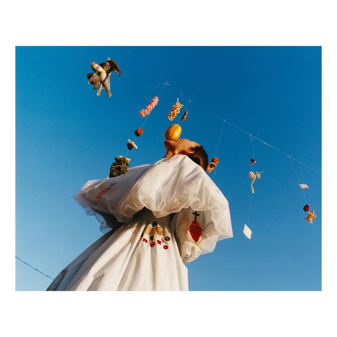 Low angle shot of a person in white dress with embroidered details, looking up at hanging objects against a clear blue sky.