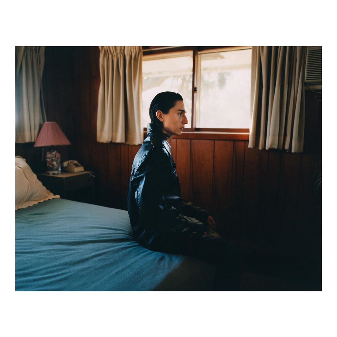 Portrait of a young adult wearing a black leather jacket sitting on a bed in a rustic indoor room with a window providing natural light.