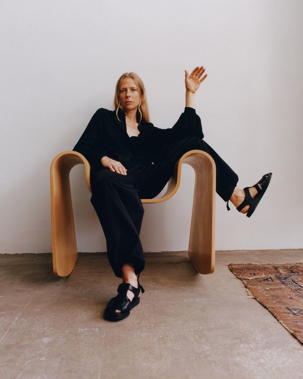 Woman with long blonde hair wearing black clothing seated in a curved wooden chair against a plain light wall.