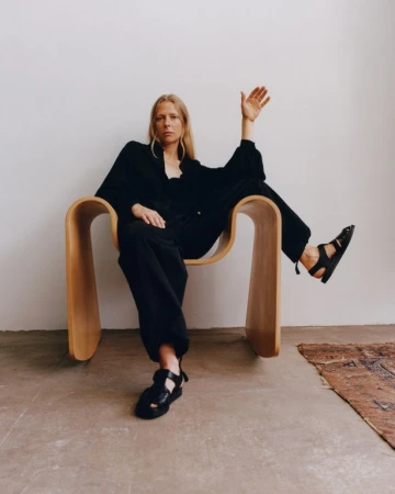 Woman with long blonde hair wearing black clothing seated in a curved wooden chair against a plain light wall.