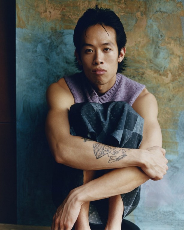 Portrait of a young adult Asian man with a neutral expression, wearing a lavender sweater and patterned shorts, seated indoors with a textured backdrop.