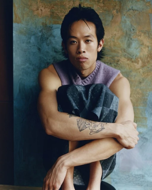 Portrait of a young adult Asian man with a neutral expression, wearing a lavender sweater and patterned shorts, seated indoors with a textured backdrop.