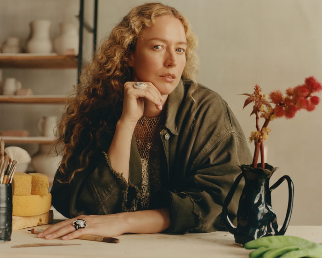 Adult woman with curly blonde hair sits at a table with pottery, paintbrushes, and flowers in a studio with natural lighting.