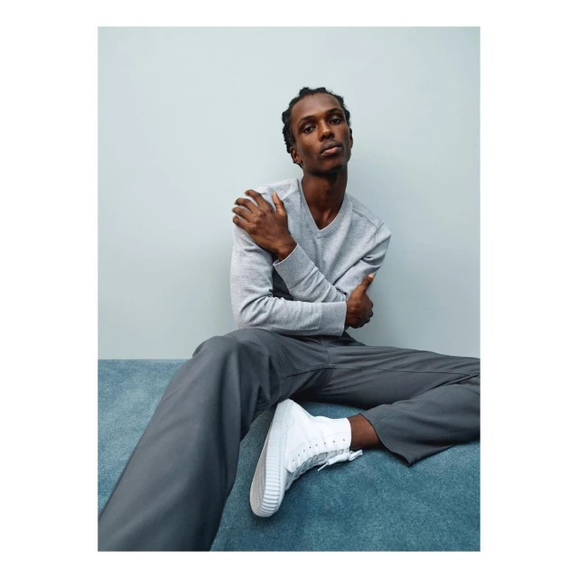 Man in grey shirt and pants sitting on teal surface against a light grey wall, wearing white high-top sneakers in studio lighting.