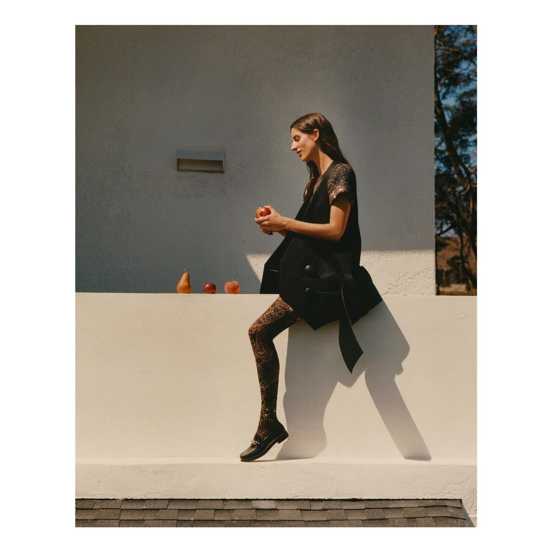 Woman in dark sleeveless coat and patterned tights holding an apple while seated on a white ledge outdoors.