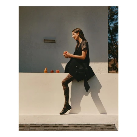 Woman in dark sleeveless coat and patterned tights holding an apple while seated on a white ledge outdoors.