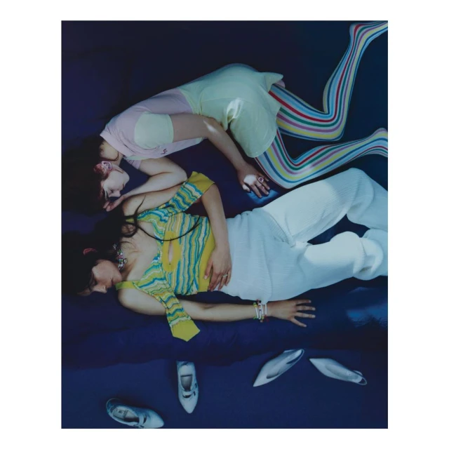 Two women in stylish attire lie on a blue surface surrounded by shoes. One wears colorful leggings and a yellow skirt, the other, white pants.