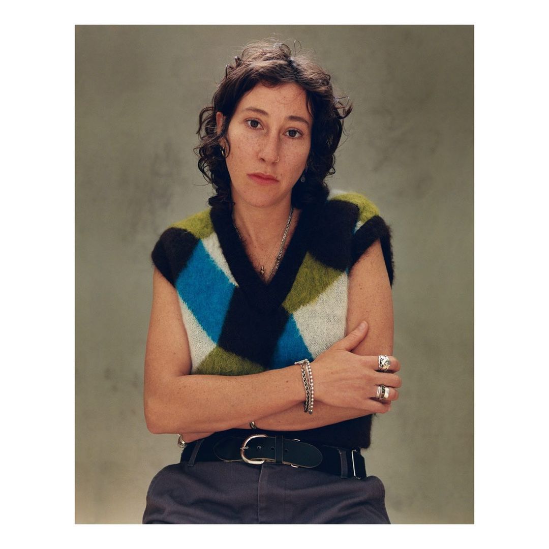 Portrait of a young woman with dark hair and a colorful argyle sweater vest, posing indoors with arms crossed against a neutral backdrop.