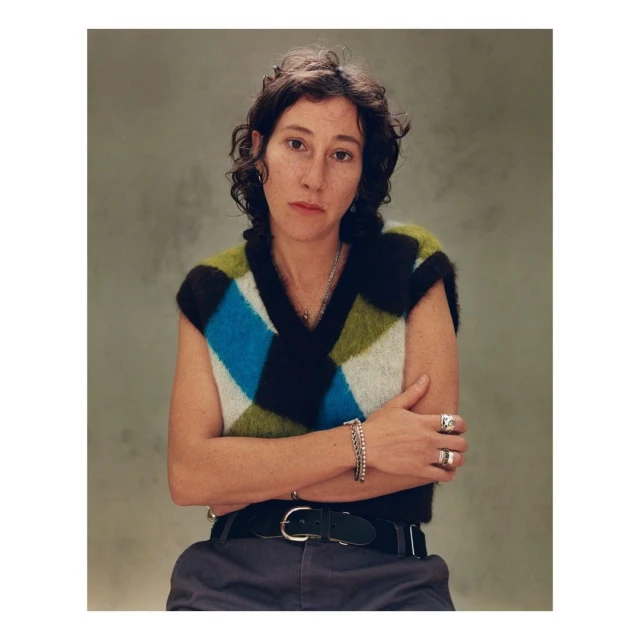 Portrait of a young woman with dark hair and a colorful argyle sweater vest, posing indoors with arms crossed against a neutral backdrop.