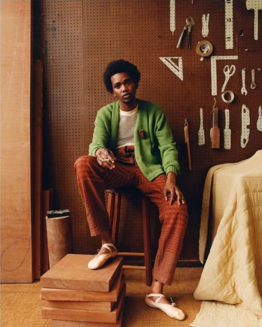 Portrait of a Black adult male seated on a wooden stool, wearing a green cardigan, plaid pants, and ballet shoes in a wood shop.