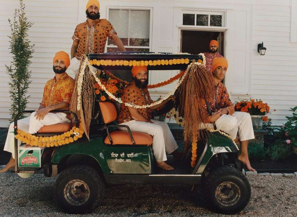 Five adult males in turbans and patterned shirts ride in a flower-decorated golf cart near a white house on a sunny day, outdoor.
