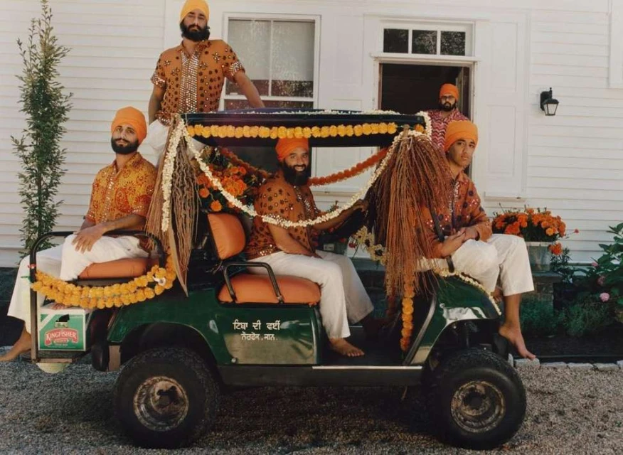 Five adult males in turbans and patterned shirts ride in a flower-decorated golf cart near a white house on a sunny day, outdoor.