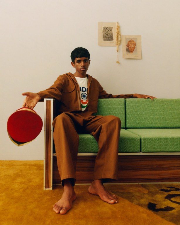 Teenager poses barefoot on green mid-century sofa in tan jumpsuit, holding red hat. Art and dried flowers hang on the wall.