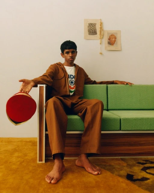 Teenager poses barefoot on green mid-century sofa in tan jumpsuit, holding red hat. Art and dried flowers hang on the wall.