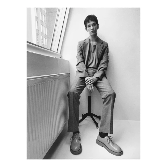 Young man in a suit poses while sitting on a chair indoors near a window, showcasing a contemporary and urban fashion aesthetic in black and white.