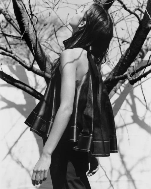 Black and white portrait of a woman in a stylish black leather top looking up at a tree branch with a blurred background.