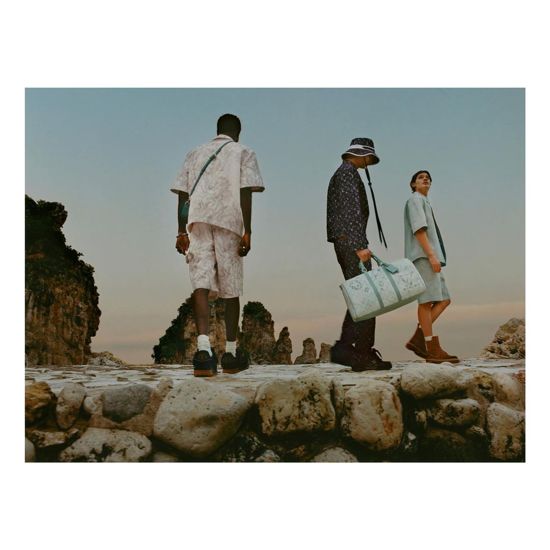 Three adult models walk along a stone path next to the ocean with rock formations, wearing casual and fashionable attire, photographed at eye-level.