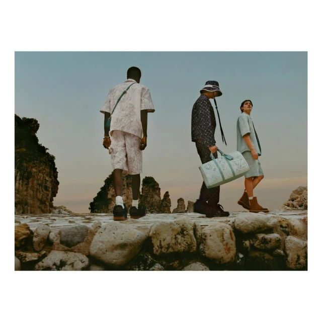 Three adult models walk along a stone path next to the ocean with rock formations, wearing casual and fashionable attire, photographed at eye-level.