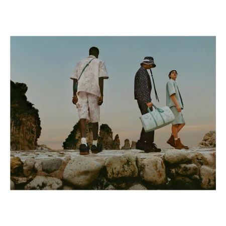 Three adult models walk along a stone path next to the ocean with rock formations, wearing casual and fashionable attire, photographed at eye-level.