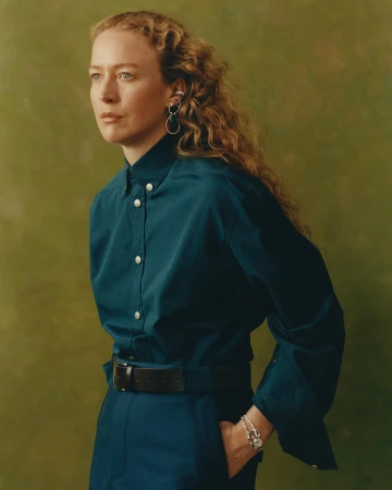 Portrait of a woman with long curly hair wearing a deep teal shirt and black belt against an olive textured backdrop