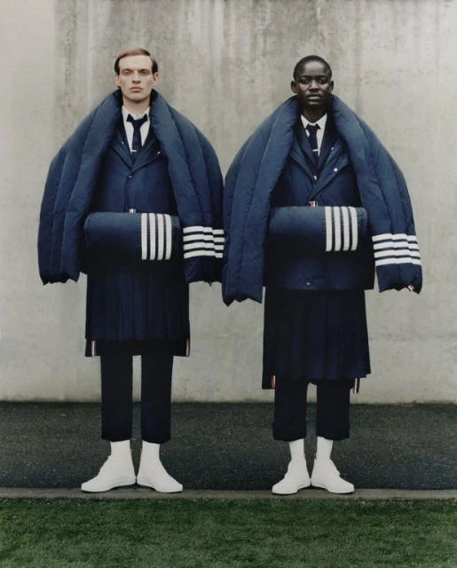 Two models in matching navy Thom Browne outfits, each with padded blazers, pleated skirts, and three-stripe accessories, stand outdoors.