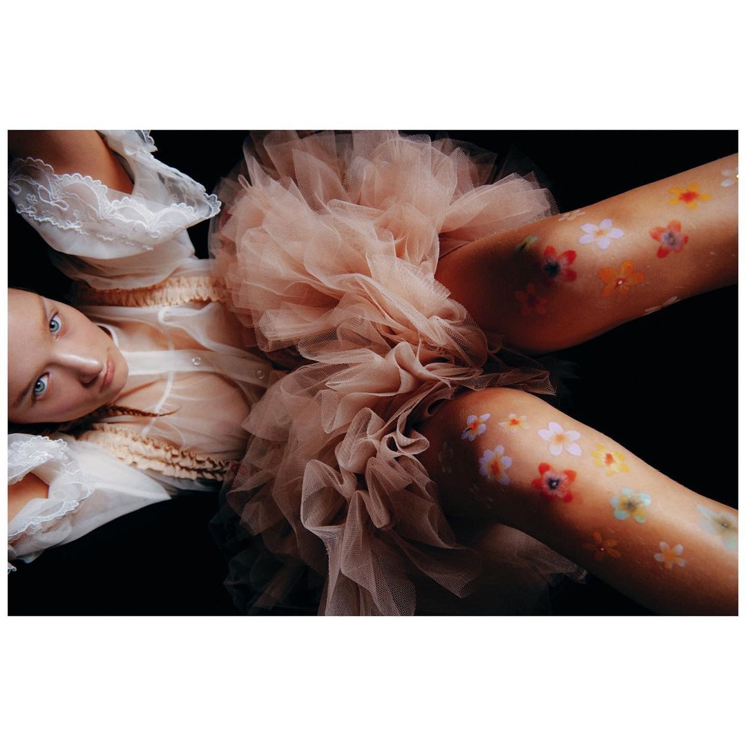 Overhead shot of a woman in a tulle dress with flower body art lying against a black background, creating a surreal and visually striking image.