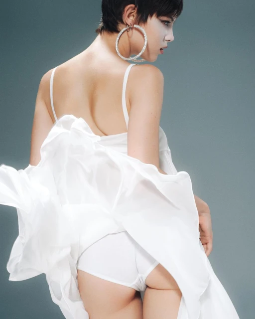 Woman with short dark hair wearing a white dress and large hoop earrings poses against a gray backdrop in a fashion studio shot.