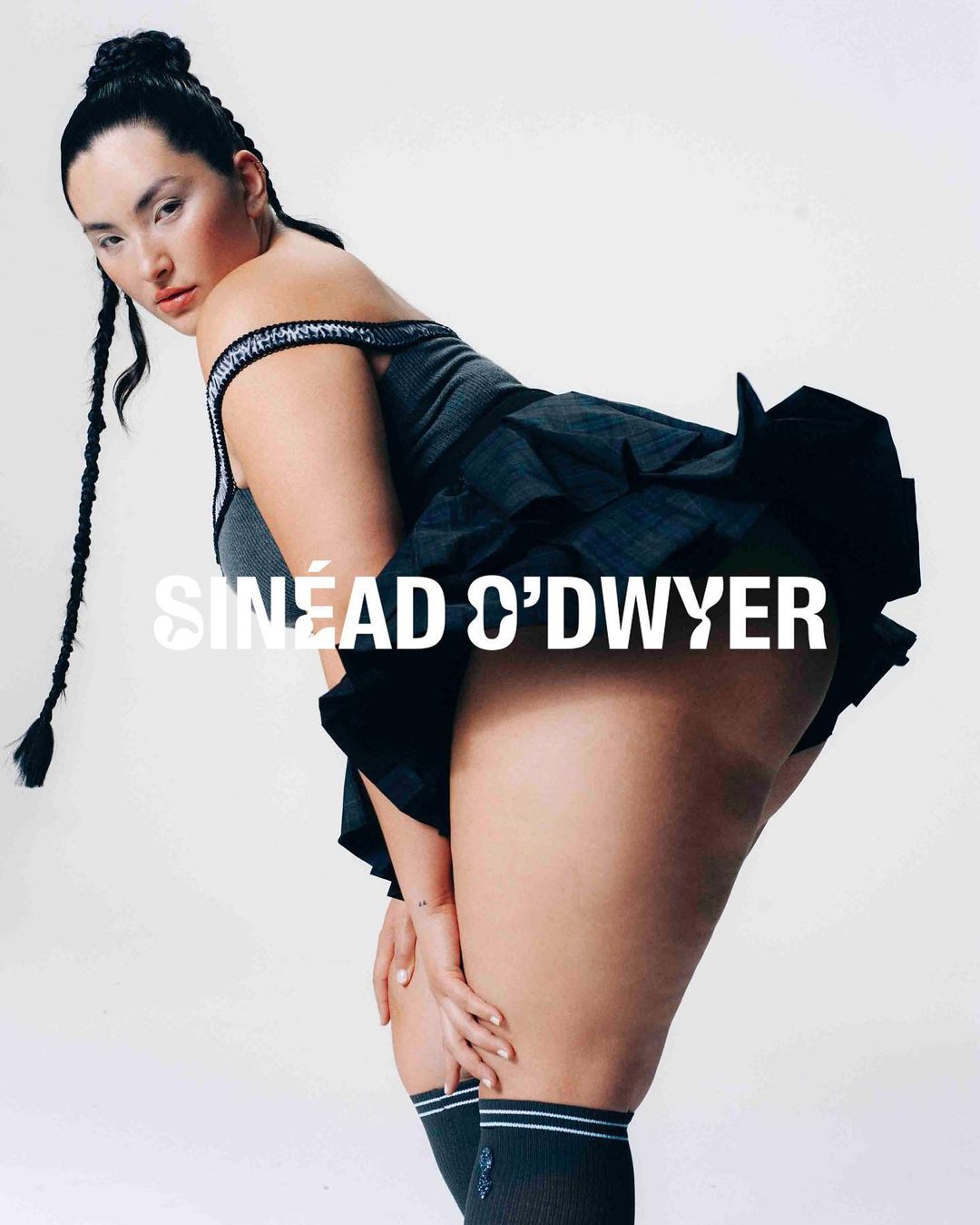 Attractive woman poses indoors, wearing gray top, black skirt, socks with white stripes, and braids. White studio backdrop.