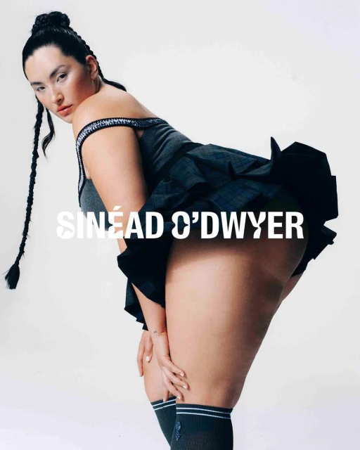 Attractive woman poses indoors, wearing gray top, black skirt, socks with white stripes, and braids. White studio backdrop.