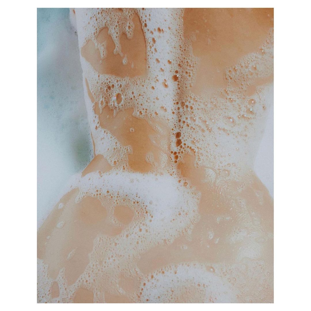 Close-up shot of an adult female's back covered in white soap bubbles in a light peach-colored bath. 