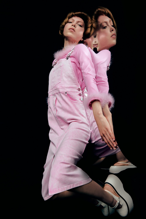 A model wears a pink overall outfit and white platform shoes, posing in front of a black backdrop with a mirrored double exposure effect.