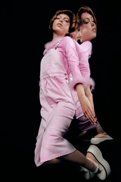 A model wears a pink overall outfit and white platform shoes, posing in front of a black backdrop with a mirrored double exposure effect.