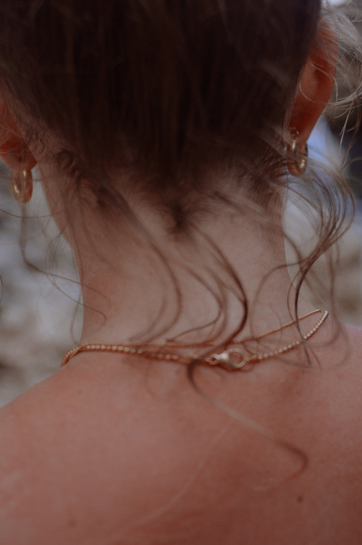 Close-up from behind of an adult female wearing a gold necklace and hoop earrings, with soft lighting and focus on the jewelry details.