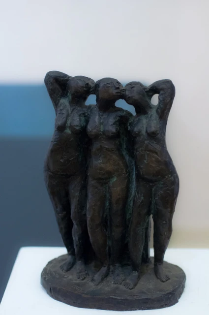 Bronze sculpture of three nude female figures with hands on their heads against a neutral studio backdrop. Fine art photography.