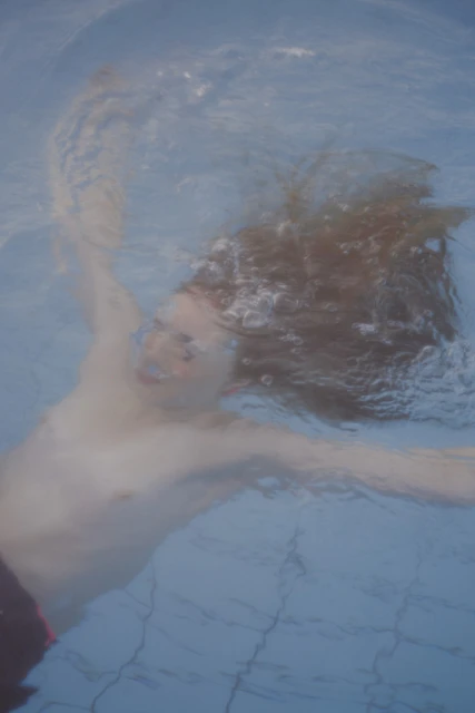Underwater view of a shirtless adult male with long hair swimming in a blue-tiled pool with sunlight reflecting on the surface.
