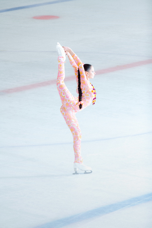 Figure skater performs a Biellmann spin on an ice rink, wearing a pink patterned catsuit, displaying flexibility and skill in an indoor setting.