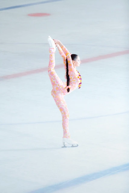 Figure skater performs a Biellmann spin on an ice rink, wearing a pink patterned catsuit, displaying flexibility and skill in an indoor setting.